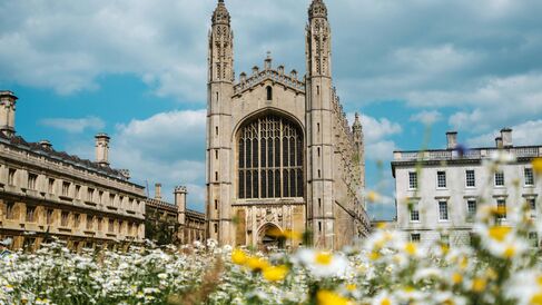 The impressive Gothic facade of King’s College Chapel, Cambridge, under blue skies and clouds. It is surrounded by other impressive Gothic buildings, flowers and daisies are seen in the foreground.