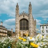The impressive Gothic facade of King’s College Chapel, Cambridge, under blue skies and clouds. It is surrounded by other impressive Gothic buildings, flowers and daisies are seen in the foreground.